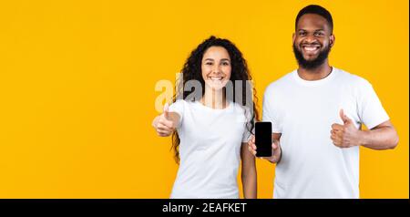 Multiethnic Couple Showing Cellphone Screen Gesturing Thumbs-Up Over Yellow Background Stock Photo