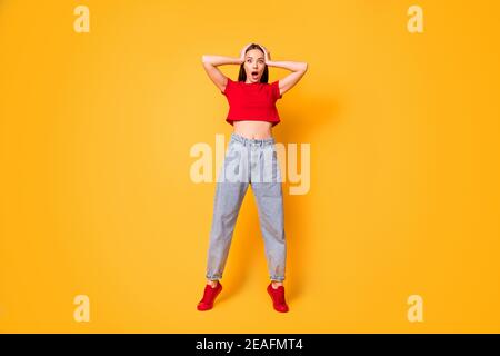 Full size photo of shocked lady hands on head stand on toes wear red crop top jeans shoes isolated on yellow color background Stock Photo