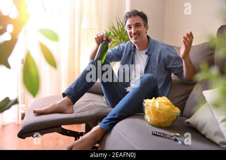 Man smiling drinking a beer from a bottle and having snack sitting on the sofa in the living room at home Stock Photo
