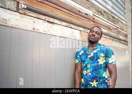 Young confident African-American male in a blue shirt leaning on a wall and gently smiling Stock Photo