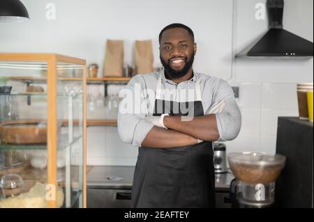 Cheerful and friendly African-American small business owner, cafe manager, bakery staff stands in confident pose with arms crossed and looks at the Stock Photo