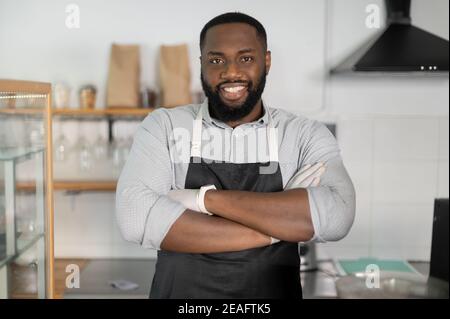 Close-up portrait of an African-American waiter standing with arms crossed in cozy loft coffee shop. A confident multiracial small business owner Stock Photo