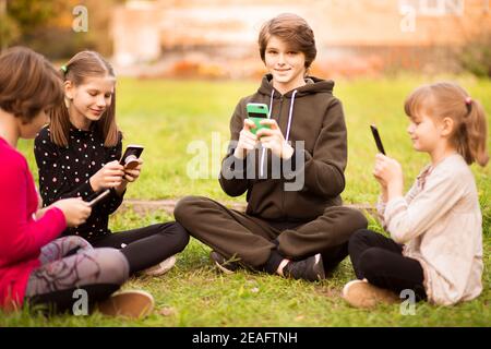 Group of busy kids looking at their phones texting sms and play games sitting with cross legs on grass outside in summer park. Selective focus Stock Photo