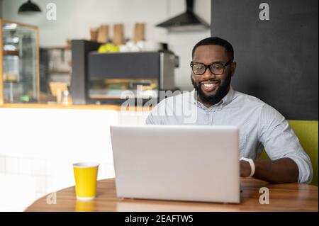 Smiling African-American guy is using a laptop for remote work sitting in the cozy cafe. A multiracial freelancer typing on the keyboard, programmer Stock Photo