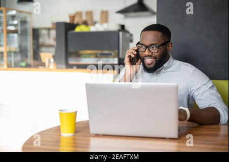 Smiling African-American guy is using a laptop for remote work sitting in the cozy cafe. A multiracial freelancer is talking on the smartphone sitting Stock Photo