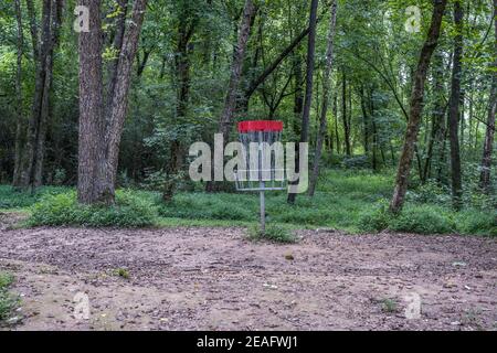 Red disc golf basket with chains in a rustic setting in the forest on a course in the shade at a park in summertime Stock Photo