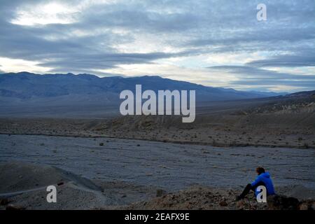 one person sitting on a stone in the evening sky with the sun breaking through clouds with a view to the panamint range and the Death Valley Wash in t Stock Photo