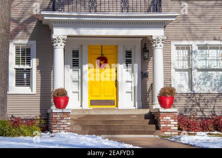 Entrance of pretty vintage house with ornate columns on porch and red valentine wreath on bright yellow door in snow Stock Photo