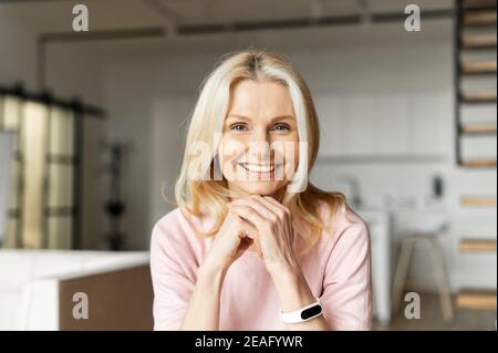 Portrait of a charming elegant mature middle-aged businesswoman with hands on a chin smiling at the camera, sitting in the modern house, wearing pink Stock Photo