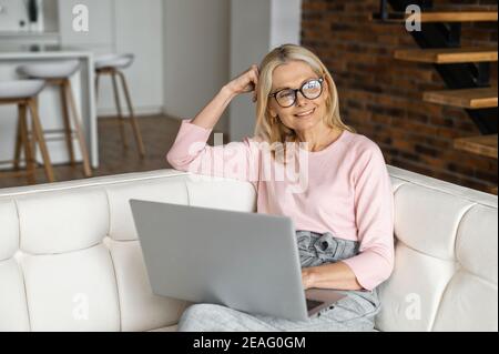 Positive charming mature blonde woman in glasses sitting on the couch, resting relaxing, thinking about items to buy, planning to watch movie online Stock Photo