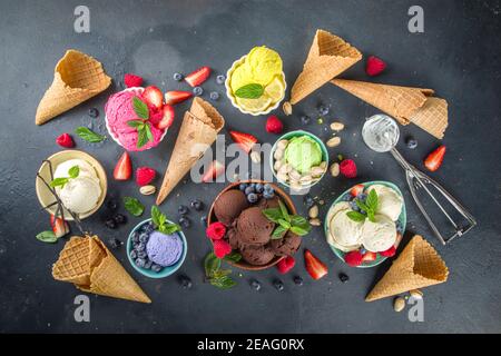 Various colorful ice cream balls in different bowls, with ice cream waffles cones and flavor ingredients - pistachio nuts, berries, lemon, chocolates, Stock Photo