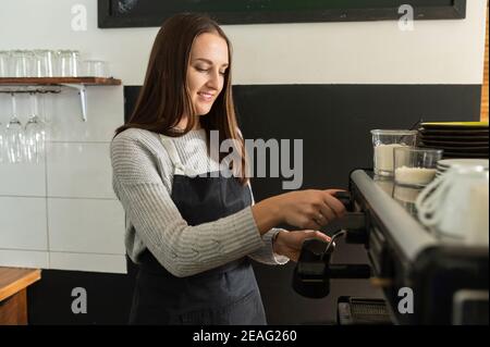 Side view an waitress wearing apron making coffee in paper cup on a professional equipment. A female barista using coffee machine in loft cafe Stock Photo
