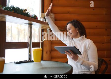 A young woman holds a tablet and waves her hand through the window sitting at the table of a cozy wooden cafe Stock Photo