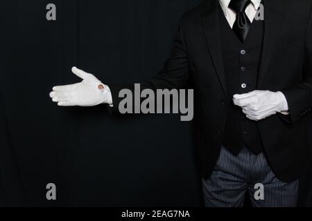 Portrait of Butler or Servant in Dark Suit and White Gloves with Welcoming Gesture. Service Industry. Professional Hospitality and Courtesy. Stock Photo
