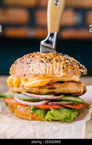Delicious juicy burger close up from Brioche Bun. Chicken steak, cheese, onion, tomato, cucumber, lettuce, stuck knife, sauce. Cafe table. Wooden boar Stock Photo
