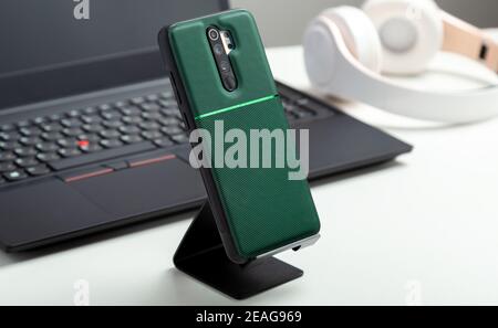 Smartphone on holder in protective case on Smartphone back near black laptop on Modern white work space. White table in home or office with phone Stock Photo