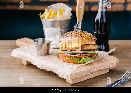 Delicious juicy burger from Brioche Bun. Chicken steak, cheese, onion, tomato, cucumber, lettuce, stuck knife, sauce, cola and fries. Cafe table. Wood