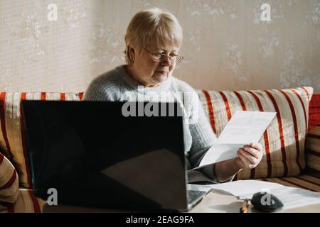 Senior woman using laptop and holding documents at home. Focused mature aged female using computer working with finance bank loan or mortgage Stock Photo