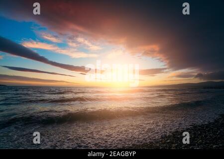 Dramatic colorful sunset on sea coast above water surface. Ocean waves splashes at beach coastline. Beautiful seascape and freedom concept. Stock Photo
