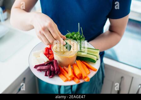 Top view close up man's hand dipping cucumber stick in hummus on the kitchen. Hummus served with raw vegetables on the plate. Healthy food lunch. Vege Stock Photo