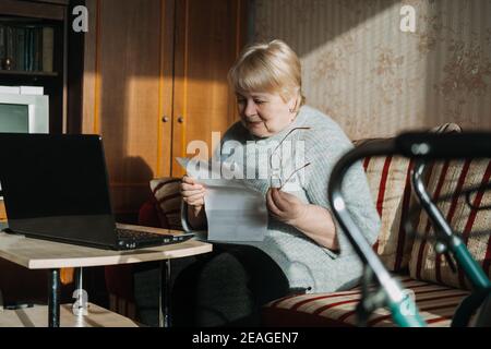 Senior woman using laptop and holding documents at home. Focused mature aged female using computer working with finance bank loan or mortgage Stock Photo