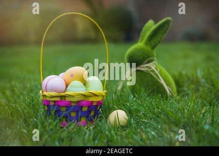 Easter basket with colored eggs and Easter bunny rabbit statuette on the green grass with dew. Easter egg hunt in the garden. Safe festive tradition d Stock Photo