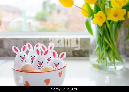 Funny Easter eggs containers with bunny ears in bowl and bouquet of yellow tulips and daffodils flower on the white kitchen table near the window. Fes Stock Photo