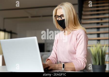 Pretty middle-aged female entrepreneur businesswoman with blond hair wearing black protective medical face mask sitting at the desk in a home office Stock Photo