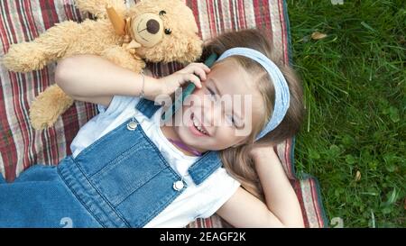 Pretty child girl laying on green lawn with her teddy bear talking on mobile phone. Stock Photo