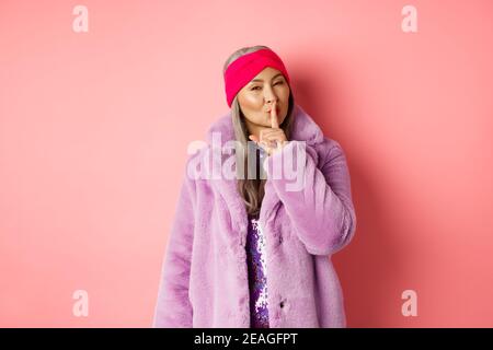 Fashion and shopping concept. Modern chinese senior woman shushing, telling a secret and looking cunning at camera, standing in trendy fake fur coat Stock Photo