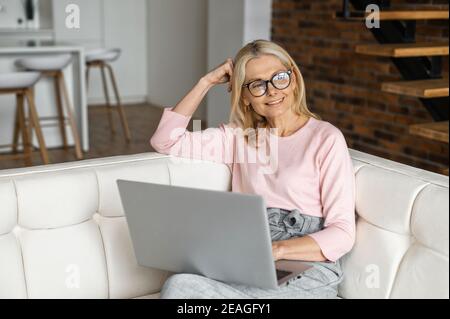 Positive charming mature blonde woman in glasses sitting on the couch, resting relaxing, thinking about items to buy, planning to watch movie online Stock Photo