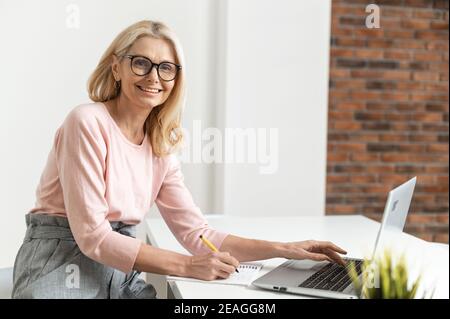 Positive charming mature blonde businesswoman in glasses sitting at the desk, typing on laptop, doing online shopping, working on a project, watching Stock Photo