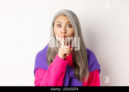 Close-up of stylish senior asian woman in hipster outfit telling hush, shushing at looking at camera, show taboo gesture, standing over white Stock Photo