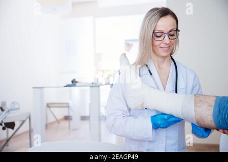 Smiling proffesional nurse in glasses bandages the leg. Stock Photo