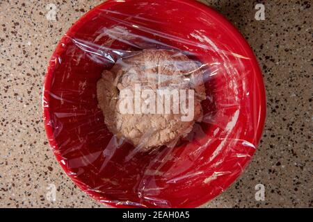 A ball of bread dough in a red mixing bowl covered with plastic wrap. Series step-by-step making homemade bread.  Frame 5 of 13 Stock Photo