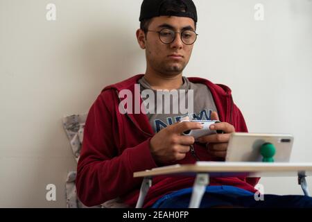 Young man wearing a red jacket and glasses playing videogames with a white controller on his smartphone at home Stock Photo