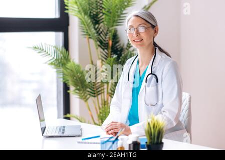 Gray-haired female middle aged general doctor dressed in medical uniform and eyeglasses looks directly at the camera at workplace at the clinic. Medical help and consultation, health concept Stock Photo
