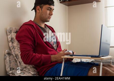 Young man wearing a red jacket playing videogames with a white controller on his blue laptop at home Stock Photo