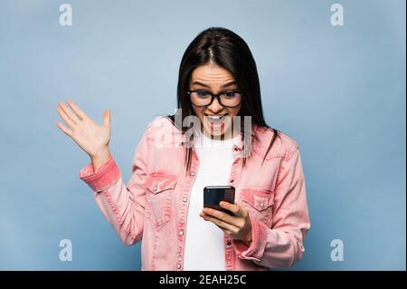 Surprised caucasian brunette girl dressed in casual stylish wear and glasses, joyfully looking at her smartphone, chatting online or getting a good news, standing against isolated blue background Stock Photo