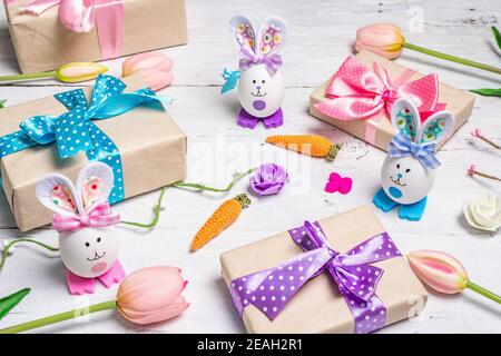 Handmade traditional Easter symbols concept. Cute rabbits from eggs, gift boxes with flowers, festive decor. White wooden background, close up Stock Photo