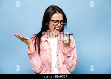 Angry emotional caucasian brunette woman shouting while talking on mobile phone with friends, family or boyfriend, standing against isolated blue background. Stock Photo