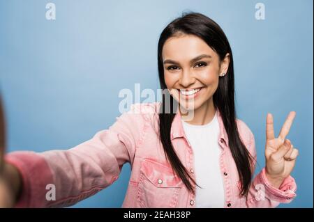 Portrait of a smiling cheerful trendy caucasian young woman dressed in stylish wear, making selfie, stands on isolated blue color background. Stock Photo