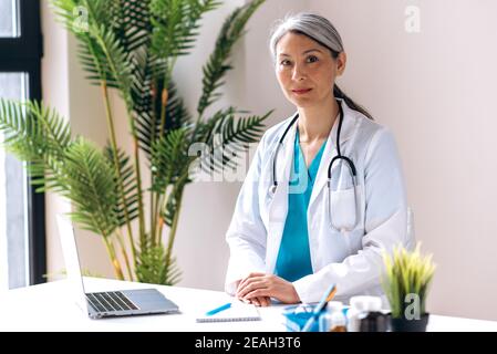 Gray-haired female middle aged general doctor dressed in medical uniform looks directly at the camera at workplace at the clinic. Doctor's consultation, health concept Stock Photo