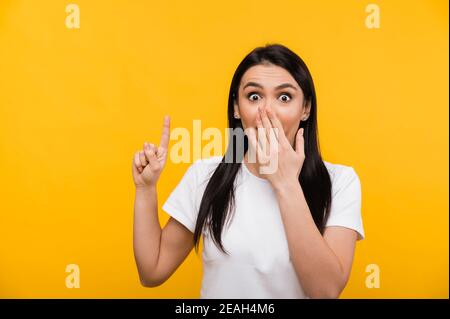 Shocked caucasian beautiful brunette woman covering her mouth with hand and surprised shows thumb up, standing against isolated orange background Stock Photo