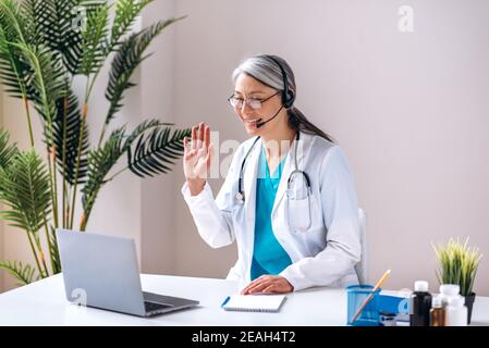 A friendly female doctor in a medical uniform and headphones consults a patient online, communicates on a video call using a laptop. Online medical assistance Stock Photo