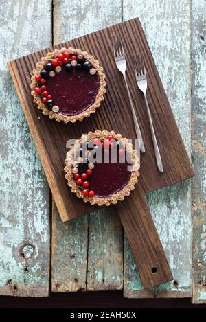 Small vegan tarts made of nuts and berry jam decorated with black, red and white currants on oak cutting board overhead view Stock Photo