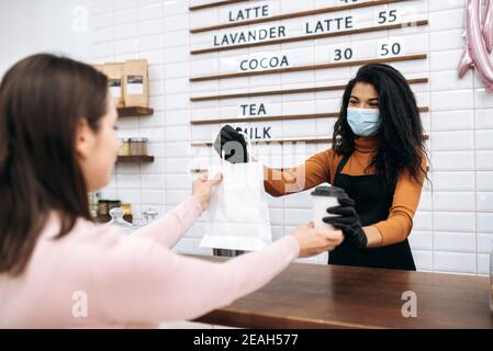 Young african american barista girl in uniform and medical mask and protective gloves gives a visitor coffee in a disposable cardboard glass and dessert in a paper bag to go. Coffee to go concept. Stock Photo