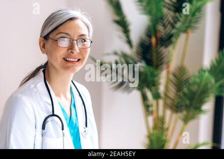 Portrait of gray-haired female middle aged doctor dressed in medical uniform and eyeglasses looks directly at the camera at workplace at the clinic and smile. Medical help and consultation, health Stock Photo