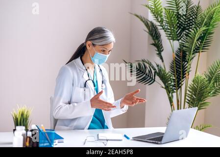 Female gray haired doctor wearing a lab coat, medical mask and headset speaking by video call using laptop with patient, online consultation. Remote medical help concept