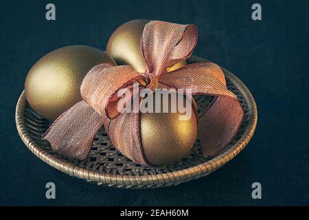 Golden Easter eggs, the front one with orange bow. Eggs are in rattan plate on dark background Stock Photo